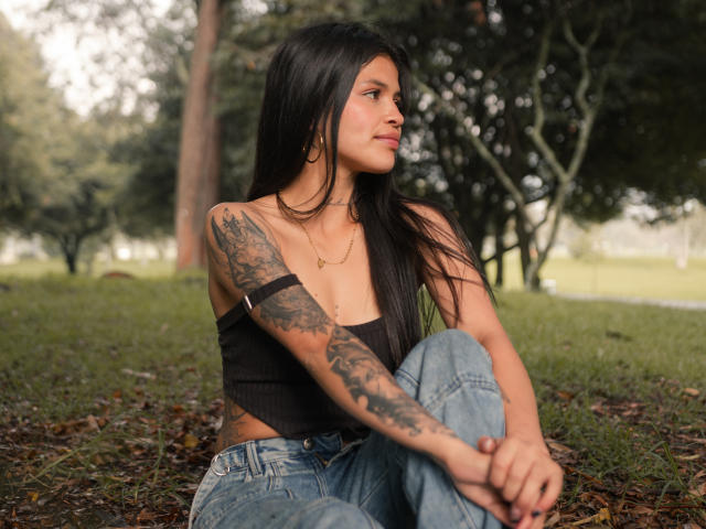 AlaiaStone with long black hair, tattooed arm, black strap top, blue jeans, sitting outside, looking to the side, small gold necklace, subtle smile, grass and trees in background.