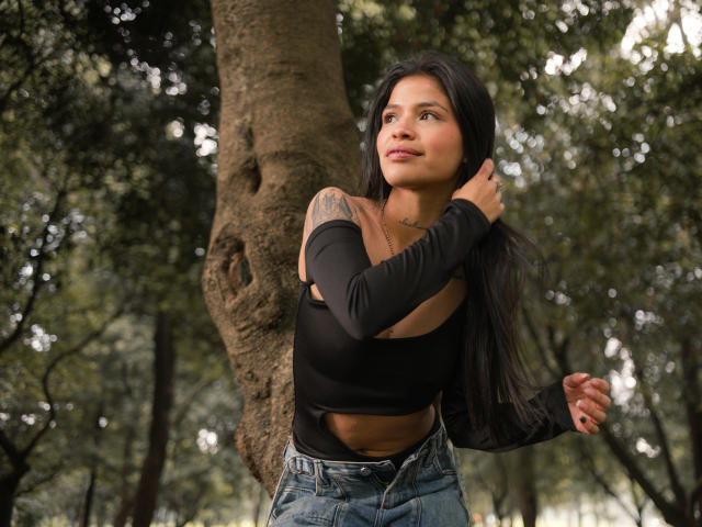 AlaiaStone, con lunghi capelli neri, indossa una canotta nera e jeans blu, è in piedi accanto a un albero, si tocca i capelli e mostra un tatuaggio sulla spalla destra..