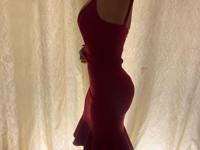 LauraVeil in a tight red dress, showing her ass and curvy figure, standing side profile, with her head out of frame. Her right arm is slightly raised.