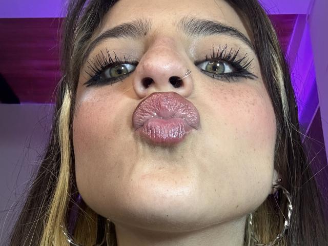 SarahhTanyas with puffy pink lips, green eyes, heavy black eyeliner, and nose piercing, blowing kiss, showing her chubby face and brown hair with blonde highlights.
