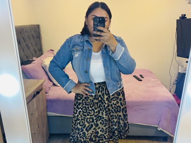 KarlaWild with hands on hips, wearing a denim jacket, white top, and leopard print skirt, taking a mirror selfie, showing her ass and medium-sized tits.