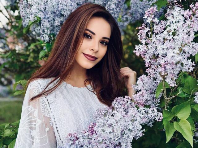 DanaSmile, con cabello castaño largo, vestido blanco de encaje y lilas morados, sonríe suavemente a la cámara. Sus senos son pequeños y está rodeada de exuberantes hojas verdes..