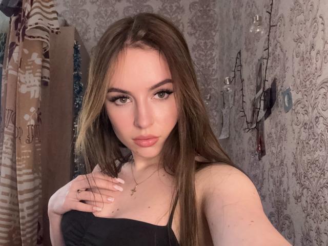 SweetSexyRosie met lang bruin haar, groene ogen en roze lippen, gekleed in een zwart topje met bandjes, waarbij haar kleine borsten zichtbaar zijn, met een hand op haar borst, subtiele make-up en een gouden kruis ketting..