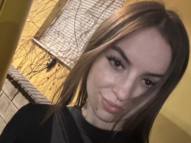 SweetSexyRosie con lunghi capelli castani, eyeliner nero, pelle chiara, un sorriso delicato e labbra rosa lucide, indossa una maglietta nera, in una stanza con pareti gialle..