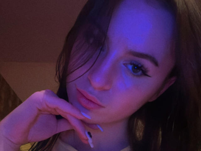 SweetSexyRosie met een paars-blauwe huid, lang bruin haar en roze lippen, die haar kin aanraakt met haar verzorgde nagels, blauwe oogmake-up en een lichte frons..