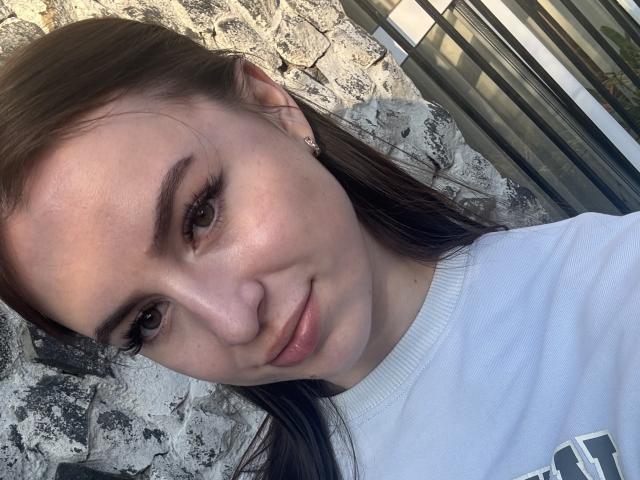 SweetSexyRosie, avec ses yeux marron et son léger sourire, porte une chemise blanche, laissant apparaître son visage lisse et ses cheveux bruns lisses, et se tient devant un mur de pierre texturé..