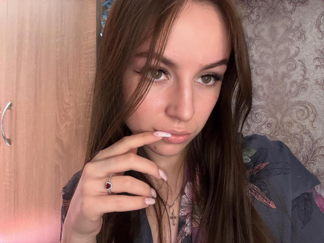 SweetSexyRosie, avec de longs cheveux bruns, des yeux verts et un vernis à ongles rose, touchant ses lèvres avec un doigt, portant une chemise bleue à motifs floraux, et montrant une bague violette..