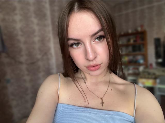 A SweetSexyRosie tem pele clara, olhos castanhos e cabelo castanho liso. Ela está vestindo uma blusa azul com alças finas, um colar delicado e olha diretamente para a câmera com uma expressão de lábios franzidos..