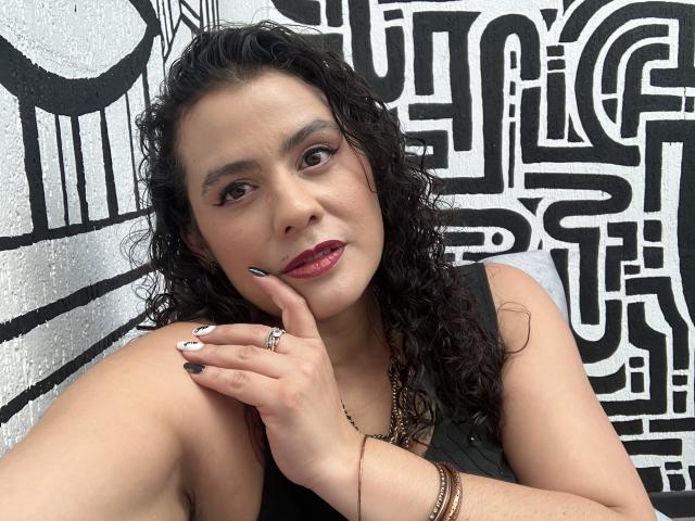 Tamarajoones, com cabelo preto encaracolado, olhos castanhos e batom vermelho, vestindo uma blusa preta, tocando sua bochecha, exibindo um anel e nail art, em frente a uma parede em preto e branco..