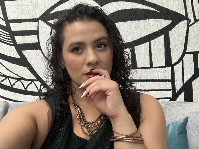 Tamarajoones, con i capelli neri ricci, rossetto scuro e una canottiera nera, che tocca le labbra con un dito, indossa diverse catene e un anello, mostrando un tatuaggio sul braccio..