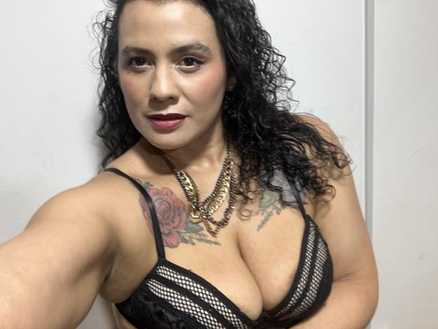 Tamarajoones met krullend zwart haar, draagt een zwarte kanten beha, toont grote borsten, heeft een tatoeage van een rode roos op haar rechter schouder, een gouden ketting en donkerrode lipstick, en glimlacht naar de camera..