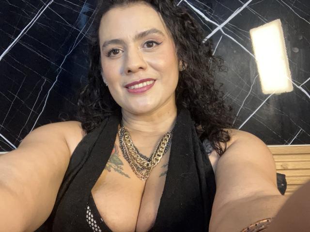 Tamarajoones, con i capelli neri ricci, sorridente, indossa un top nero a halter che mette in mostra il suo seno abbondante, una collana a catena dorata, tatuaggi sul petto e rossetto rosso..