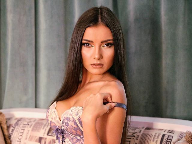 MiaAddison with long brown hair, wearing a purple lace bra, pulling the strap off her shoulder, looking at the camera with a serious expression, showing her tits.