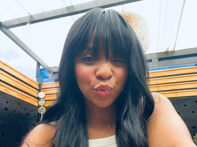 LisaMendez with long black hair, winking, and puckering her lips in a kissy face, showing her brown nose and wearing a white top, under a transparent roof.