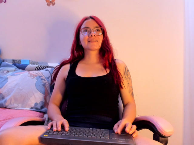 EimmyHoskX with red hair, glasses, black tank top, tattoos on arms, sitting in chair, hands on keyboard, smiling, pink armrests, blue patterned pillow in background.