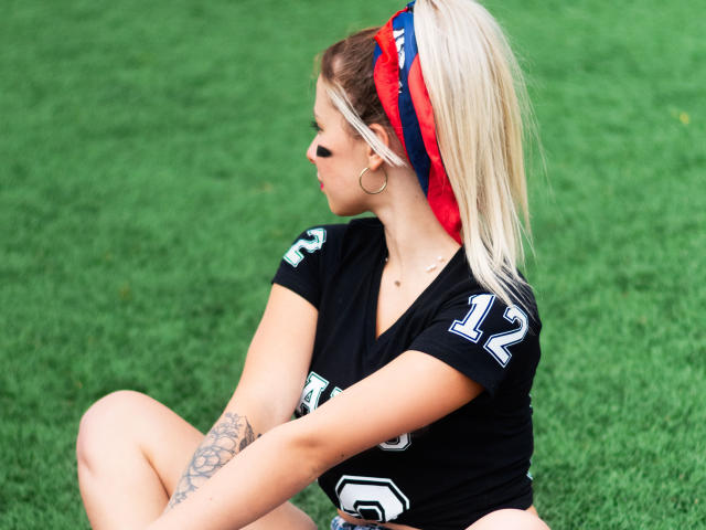 NatSorian, blonde, in black NFL jersey, number 12, sitting on grass, red and blue headband, tattoo on left arm, looking sideways, hoop earring, short black top, bare midriff.