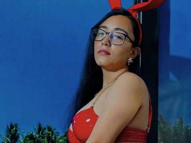 JewelryQueen con lunghi capelli neri, orecchie da coniglio rosse, occhiali, reggiseno rosso scintillante, che mostra i suoi seni, indossa una collana d'oro, in piedi davanti a uno sfondo blu..