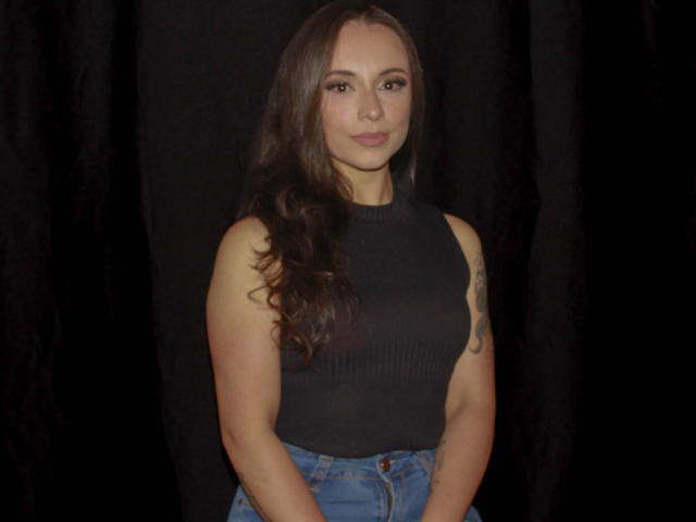 BeautyBellaX with long brown hair, black sleeveless top, and blue jeans, showing a tattoo on her right arm, standing against a black background, looking straight at the camera.