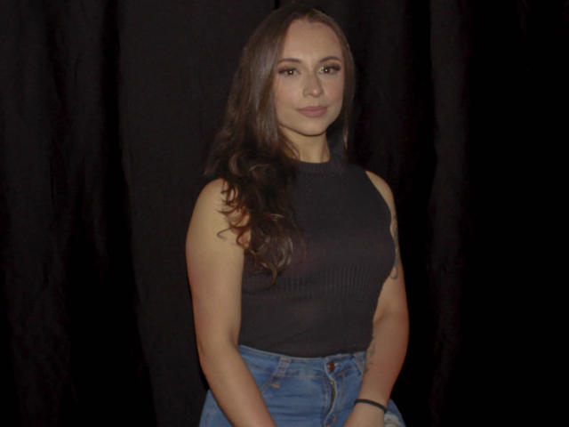 BeautyBellaX with long brown hair, wearing a black sleeveless top and blue jeans, standing against a dark background, showing a subtle smile, and a blue wristband on her right.