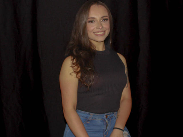 BeautyBellaX with long brown hair, smiling, wearing a black sleeveless top and blue jeans, standing against a dark background, showing a cute ass and small tits.
