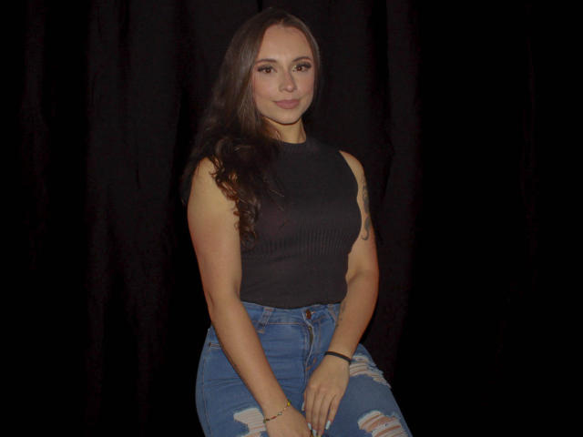 BeautyBellaX with long brown hair, black sleeveless top, and blue ripped jeans, sitting against a dark background. She has a tattoo on her right arm and a bracelet.