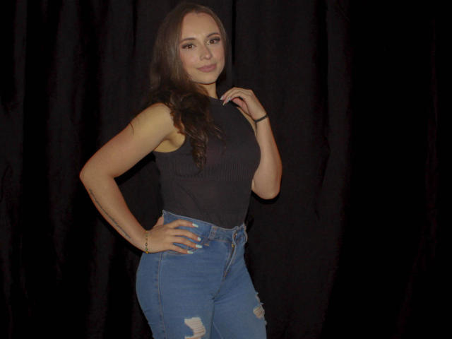 BeautyBellaX with brown hair, black sleeveless top, and blue ripped jeans, standing against a black curtain, hand on hip, smiling, showing her curvy ass and small tits.