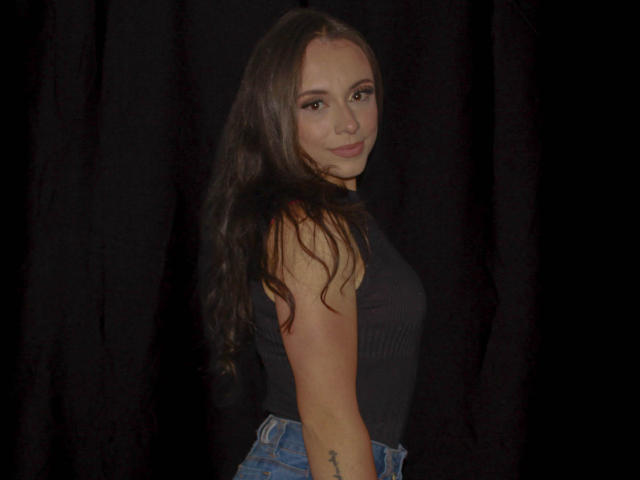 BeautyBellaX with long brown hair, wearing a black sleeveless top and blue jeans, smiling at the camera, showing a tattoo on her right arm.