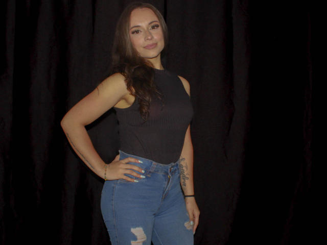 BeautyBellaX with long brown hair, black sleeveless top, and blue ripped jeans, standing with one hand on hip, showing tattoo on left forearm, smiling slightly.