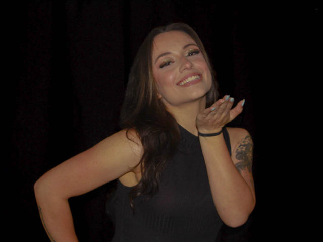 BeautyBellaX with long brown hair, smiling, wearing a black sleeveless top, tattoo on right arm, holding hand up, black background, black bracelet on right wrist.