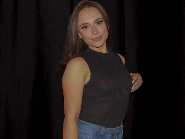 BeautyBellaX with medium brown hair, dark sleeveless top showing small tits, blue jeans, hand on chest, smiling, black curtain background, standing, photo.
