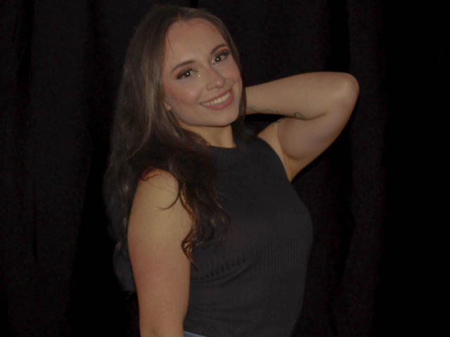 BeautyBellaX has long brown hair, smiling, wearing a black sleeveless top, with her right arm raised behind her head. She has small tits and a cute face.