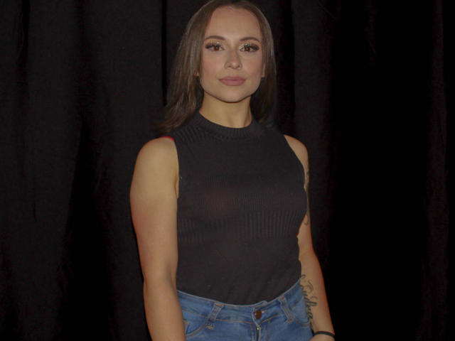 BeautyBellaX with brown hair, wearing a black sleeveless top showing her tits, and blue jeans, standing against a dark curtain, looking at the camera with a slight smile.