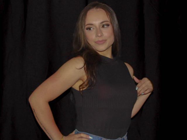 BeautyBellaX with long brown hair, wearing a sleeveless black top, smiling, hand on hip, standing against a dark curtain, showing her taut, small tits, and slim arms.