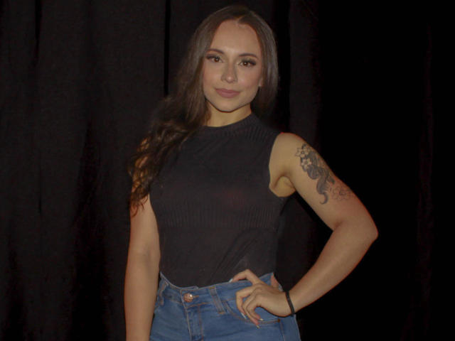 BeautyBellaX with long brown hair, black sleeveless top, blue jeans, tattoo on right arm, hand on hip, dark background, smiling subtly, no visible boobs or ass.