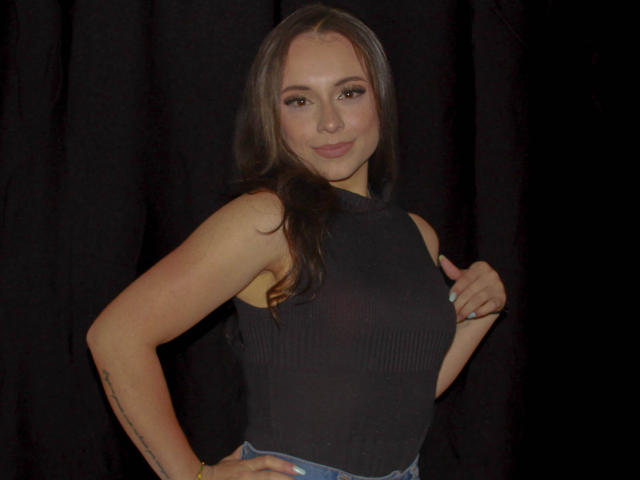 BeautyBellaX with long brown hair, wearing a black sleeveless top, smiling, hand on hip, blue jeans, tattoo on right arm, black curtain background.