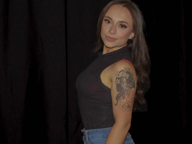 BeautyBellaX with long brown hair, wearing a black sleeveless top, showing a detailed tattoo on her right arm, and standing against a dark background. She's looking at the camera.