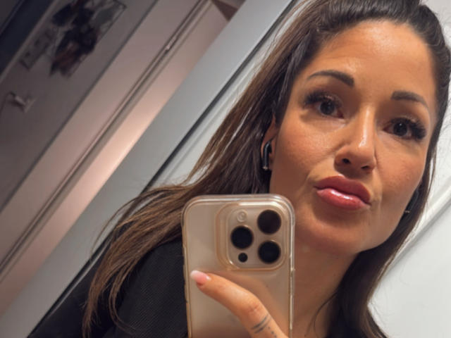 JoliSysy with glossy pink lips, brown eyes, and long brown hair, taking a selfie with a gold iPhone, holding it in front of her face, wearing a black top.