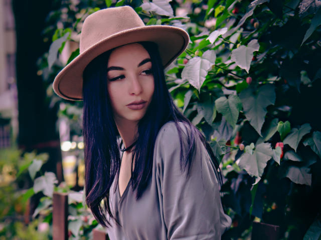 CamilleRios has dark purple hair, a beige hat, and a grey shirt, standing against leafy green vines. She's looking to the side with a slight, sexy smirk.