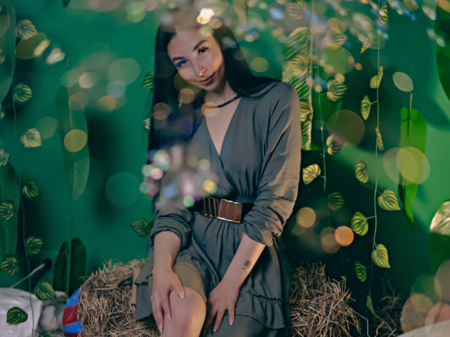 CamilleRios, with long black hair, wears a green dress with a brown belt, sitting on a hay bale, green bokeh background, looking down with a slight smile.