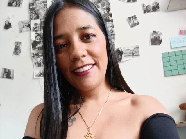 PauletteLoveX with long black hair, smiling, showing teeth, purple lipstick, gold necklace, black off-shoulder top, small tattoo on left chest, medium brown skin, cute.
