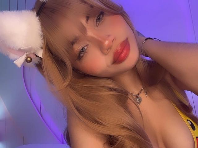 LiliMays with fluffy white cat ears, red lips, and long blonde hair, wearing a yellow top, showing her tits, and a heart necklace, under purple lighting.