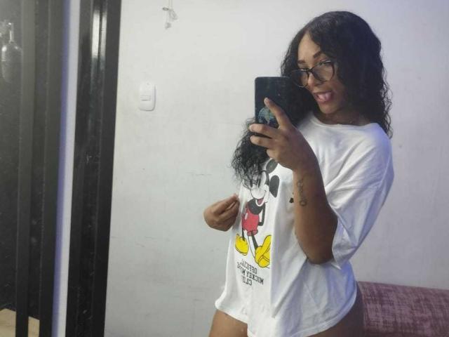 ThiainaRose, com cabelo preto cacheado, óculos e uma camiseta branca do Mickey Mouse, mostrando o bumbum, sorrindo e segurando o celular em um selfie no espelho. A tatuagem em seu braço é visível..
