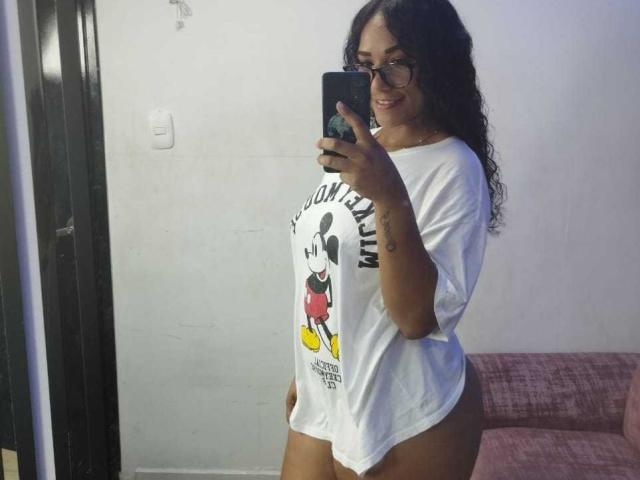 ThiainaRose, avec ses cheveux noirs bouclés, ses lunettes et son t-shirt blanc avec Mickey Mouse, montrant ses fesses, debout en profil, prenant un selfie devant un miroir, en souriant..