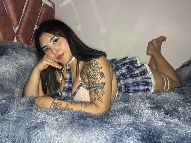 KatiaRodriguez, lying on a blue fluffy blanket, wearing a blue plaid schoolgirl outfit, has tattoos on her right arm, and is looking at the camera with a slight smile.