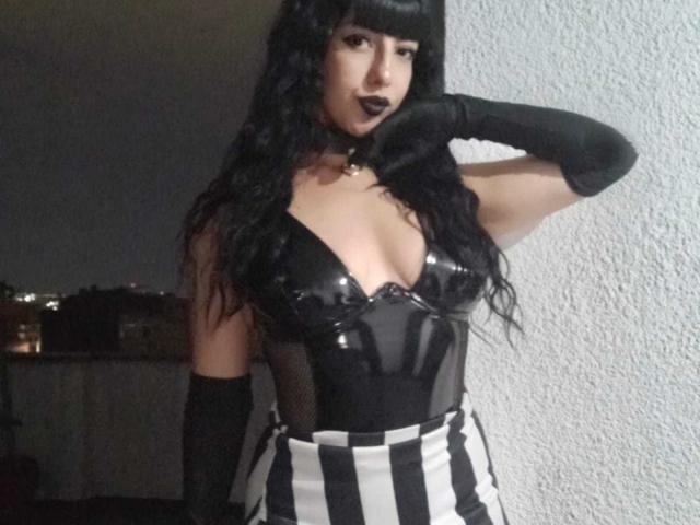 Katherin, in einem schwarzen Latexbüstier, das ihre Brüste zeigt, mit einem gestreiften Rock, schwarzen Handschuhen, dunklem Lippenstift und langen schwarzen Haaren, posiert mit einer Hand an der Wand..