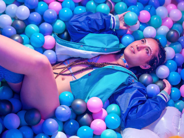 HellenK lies in a blue and pink ball pit, wearing a blue jacket and braided hair, with a choker, and her tits and ass exposed, looking seductive.