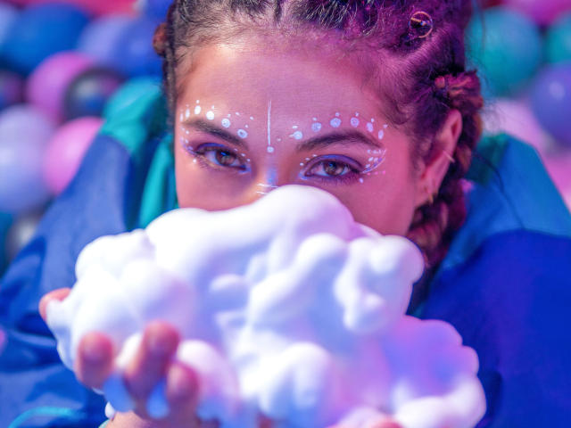 HellenK with braided red hair, blue jacket, white face paint dots, and pink highlights, eating fluffy white cotton candy, holding it near her mouth.