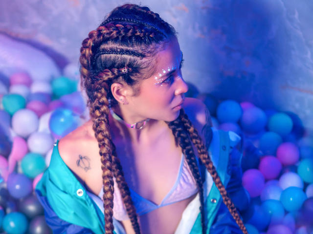 HellenK with braided brown hair, blue jacket, white bikini top, purple glitter on her face, small tattoo on left shoulder, colorful balls in background, looking to the side.
