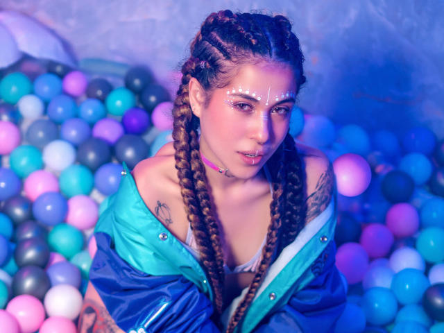 HellenK with braided brown hair, white facial markings, blue-green jacket, showing tits, tattoo on left shoulder, blue-purple ball pit background, looking at camera, slightly parted pink lips.