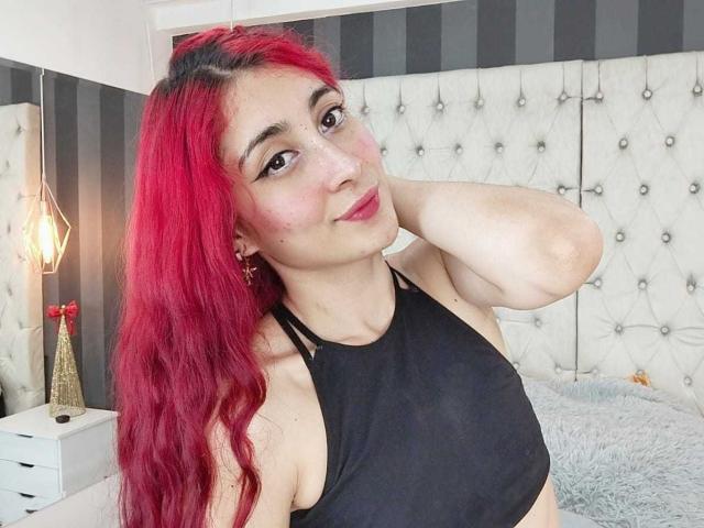 VictoriaMillar with red hair, black halter top, hand in hair, smiling, pink blush, black eyeliner, small earrings, light skin, bed with white tufted headboard, lamp in background.